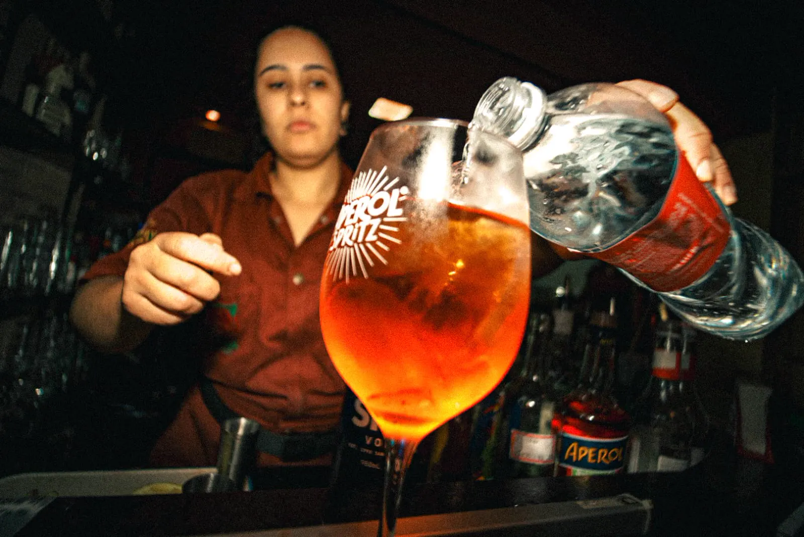 Taça de spritz servida no balcão, com a luz baixa do bar ao fundo.